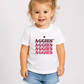 A young toddler girl with blonde curly hair in a high ponytail wearing a white t-shirt with 'AGGIES' printed in dark red and pink, paired with denim shorts, against a plain light gray background.