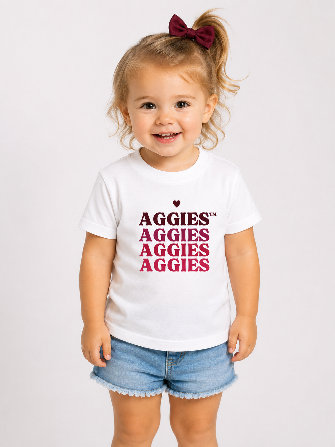 A young toddler girl with blonde curly hair in a high ponytail wearing a white t-shirt with 'AGGIES' printed in dark red and pink, paired with denim shorts, against a plain light gray background.